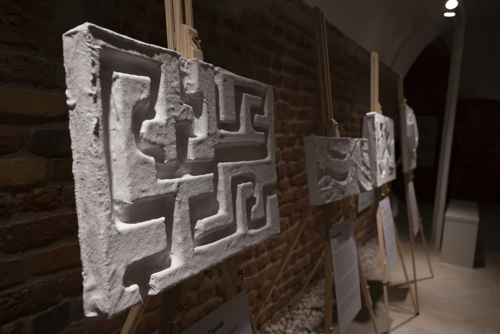 Lucrări tactile cu forme arhitecturale 3D expuse pe șevalete într-o galerie de cărămidă.