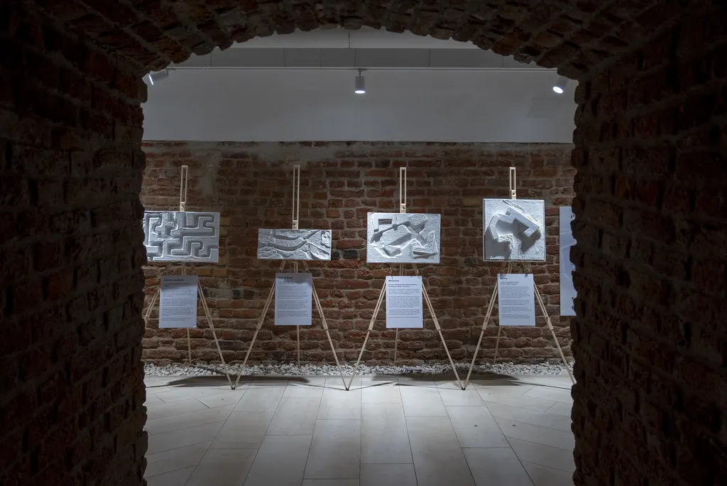 Lucrări tactile cu forme arhitecturale 3D expuse pe șevalete într-o galerie de cărămidă.